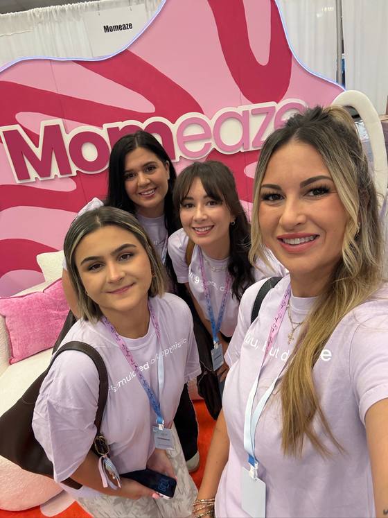 The Momeaze team in lavender tees, crowded into a selfie in front of the giant Momeaze booth sign at a conference