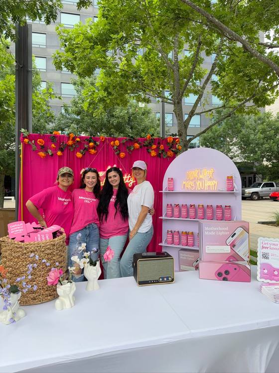 Four of the team behind the Momeaze IRL pop-up table, in hot pink tees, with a wall of Default Parent Energy koozies behind them
