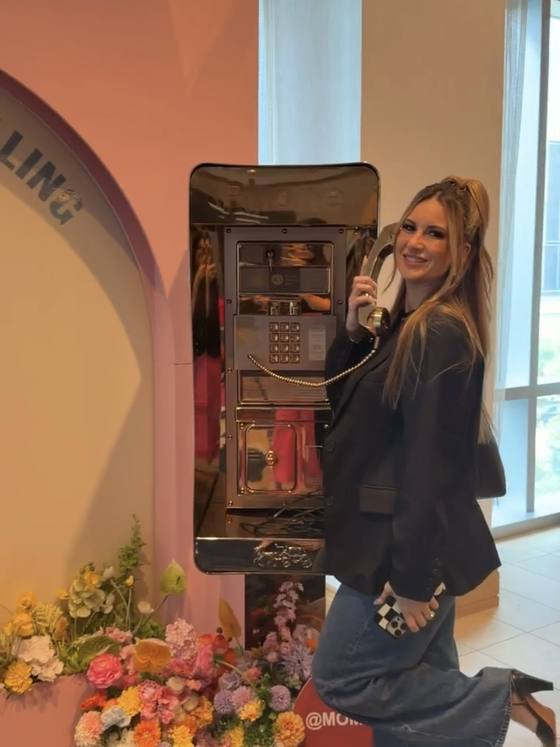Kayla Kugler at a pink payphone installation banked with flowers at Mom 2.0