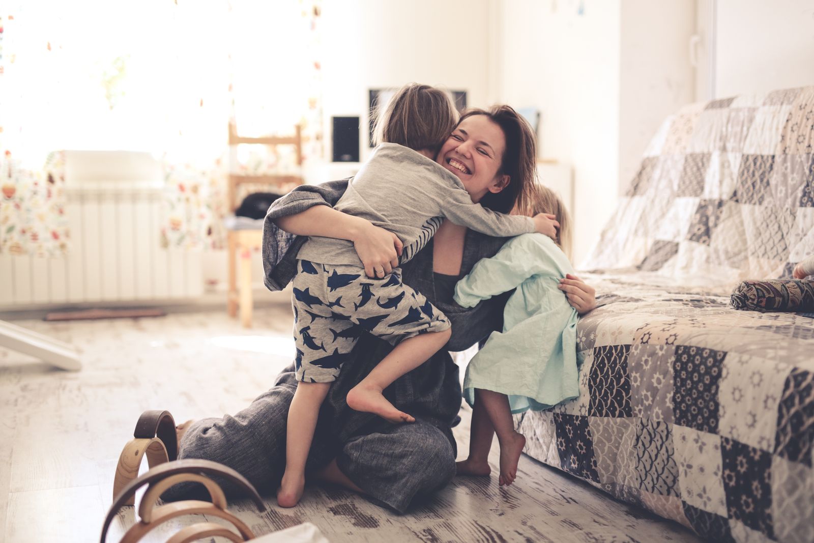 A mother sitting on the bedroom floor being hugged by her two children