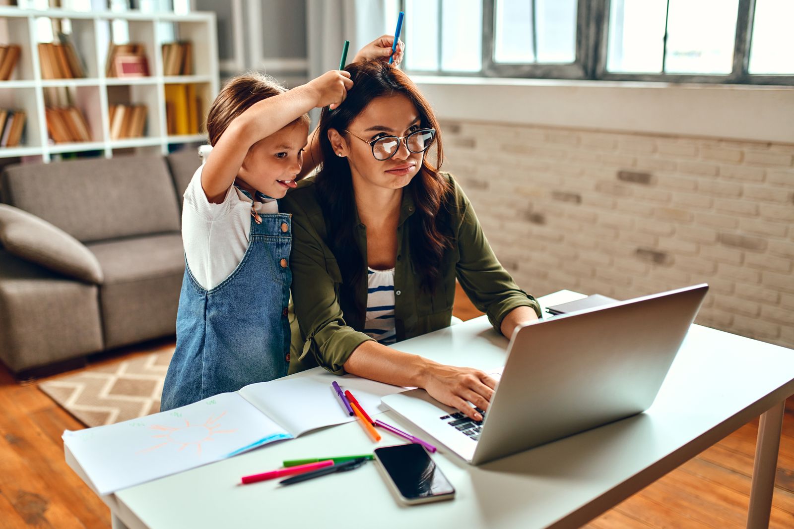 A mother working at her laptop at the dining table while her daughter leans over her, hands over her eyes