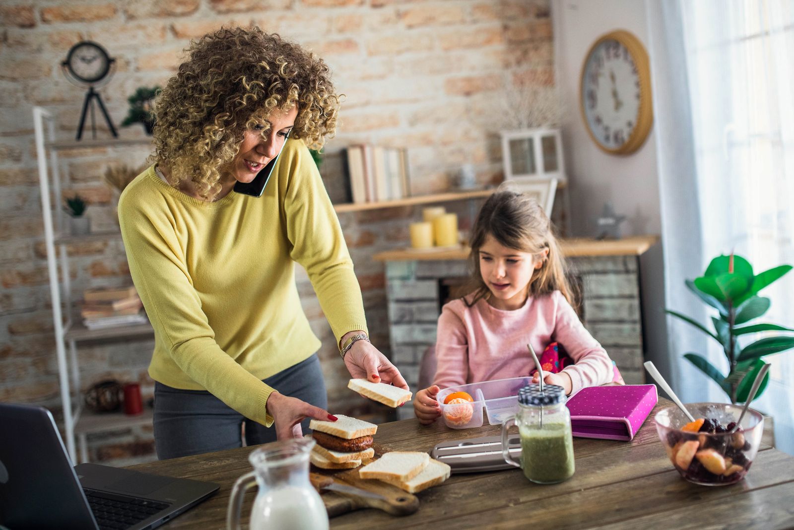 A mother making a school lunch with her daughter at the counter, phone to her ear