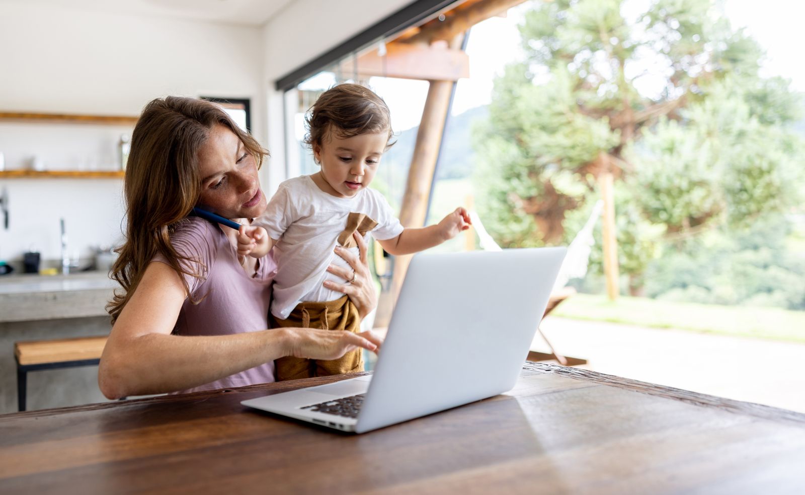 A mother holding her toddler while working at a laptop in a bright kitchen