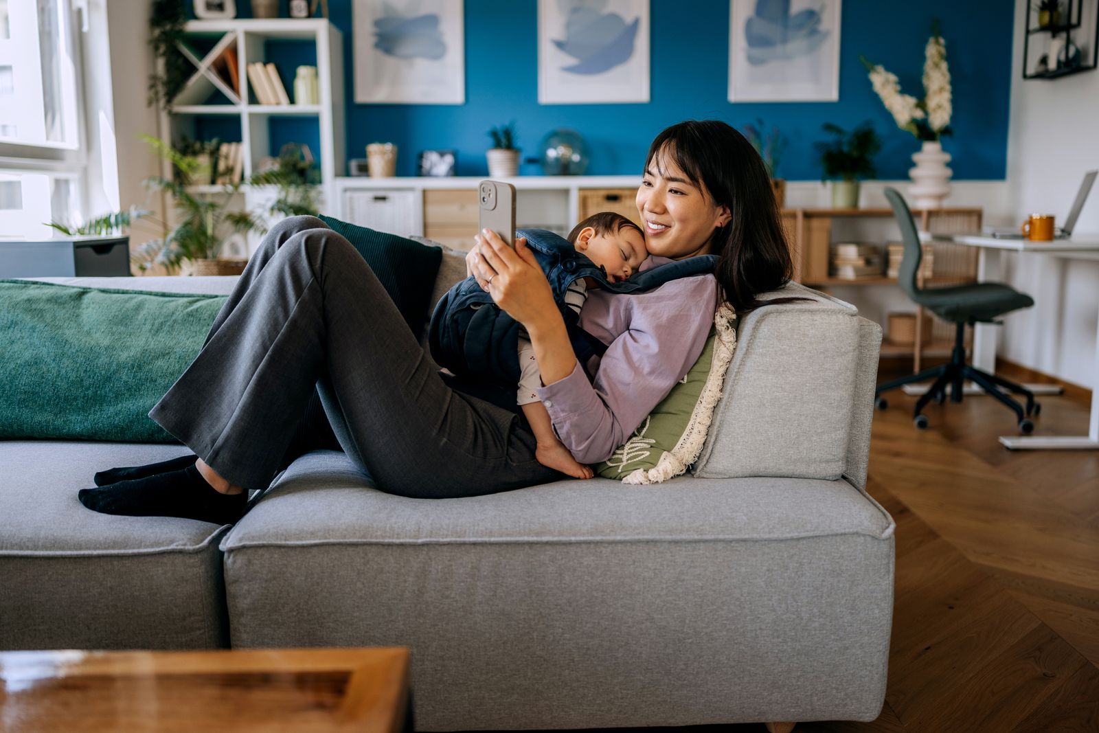 A mother resting on the sofa with her sleeping newborn against her chest, looking at her phone