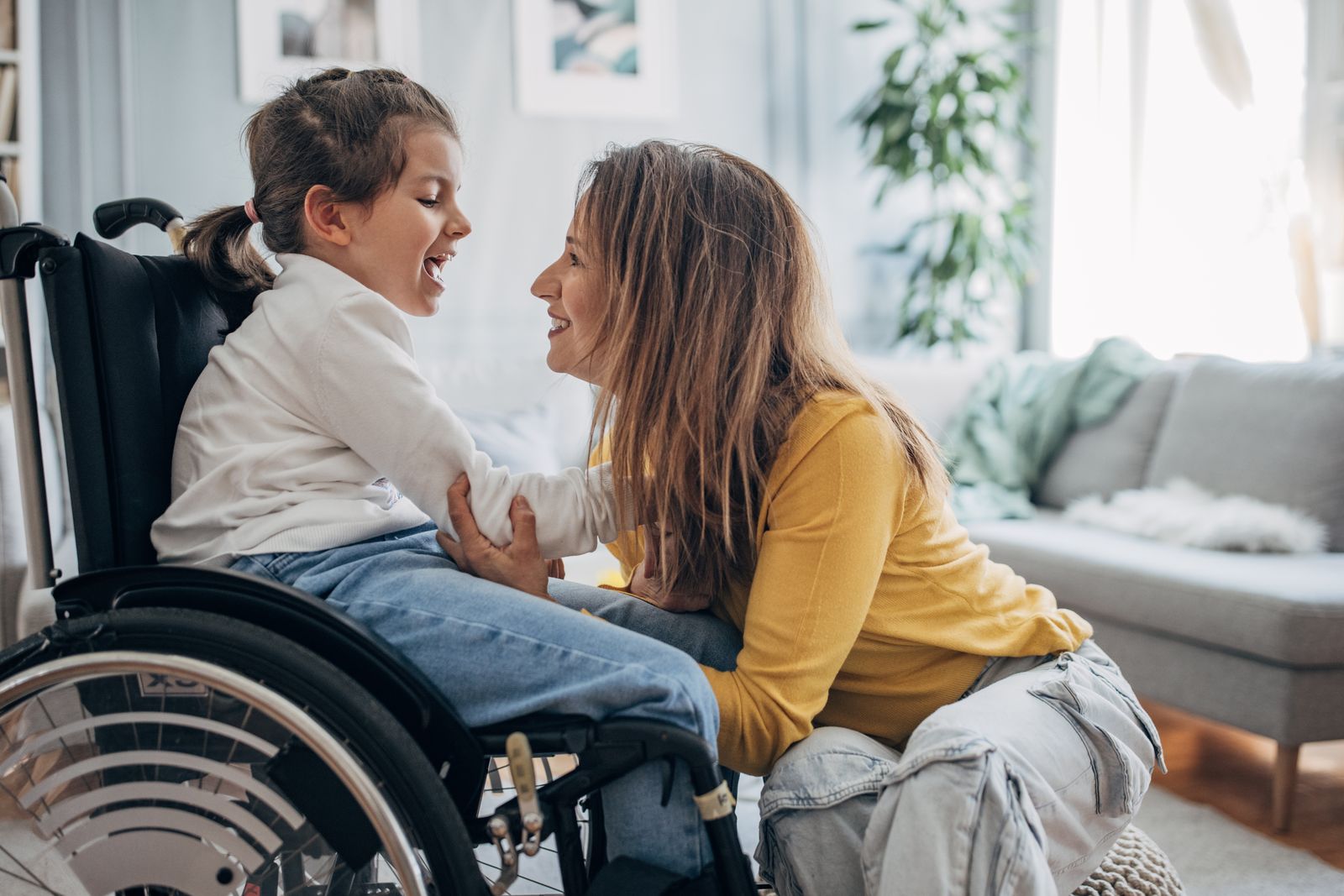 A mother and her daughter, who uses a wheelchair, laughing together in their living room