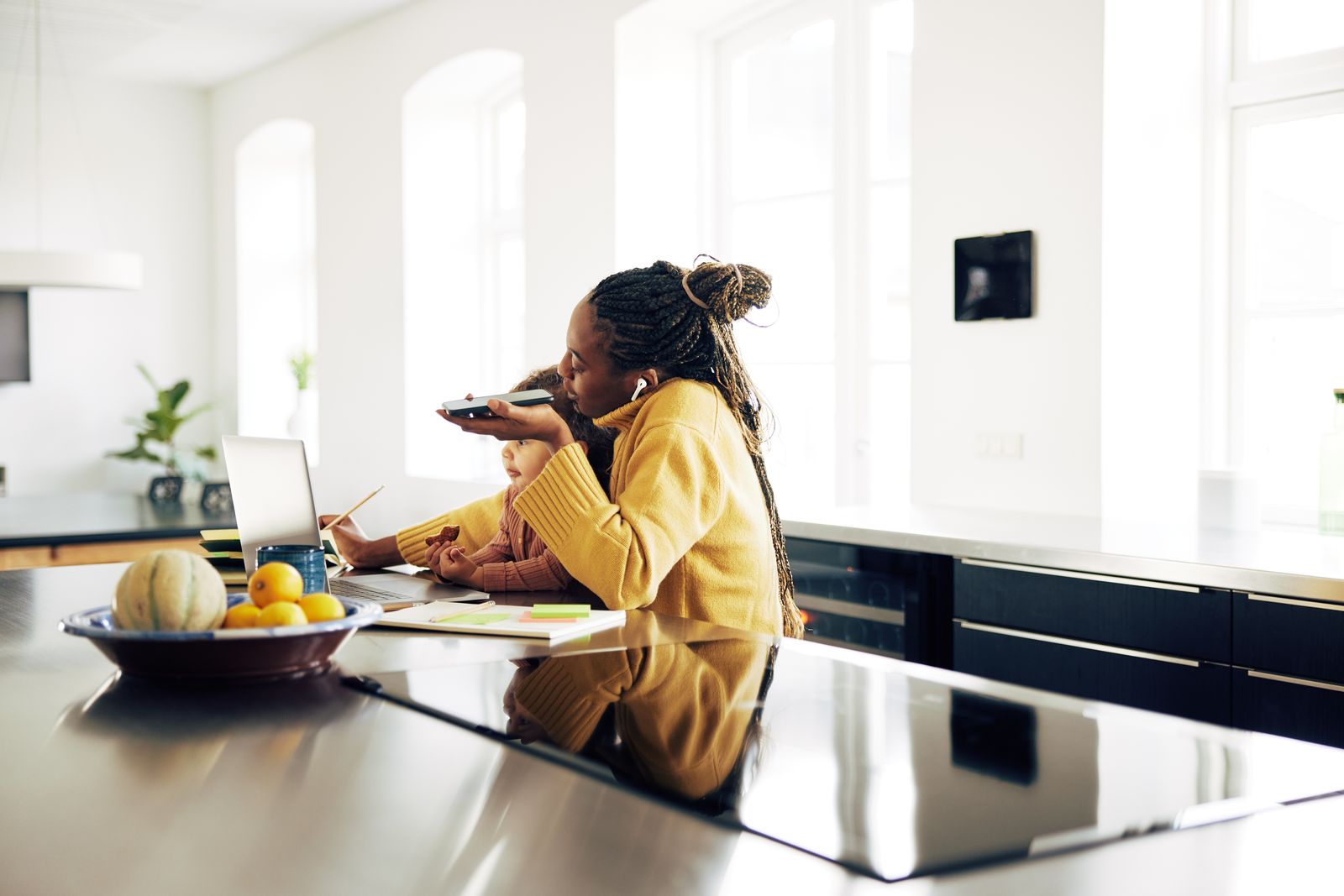 A mother working at a kitchen island with her laptop open and her child beside her, speaking into her phone