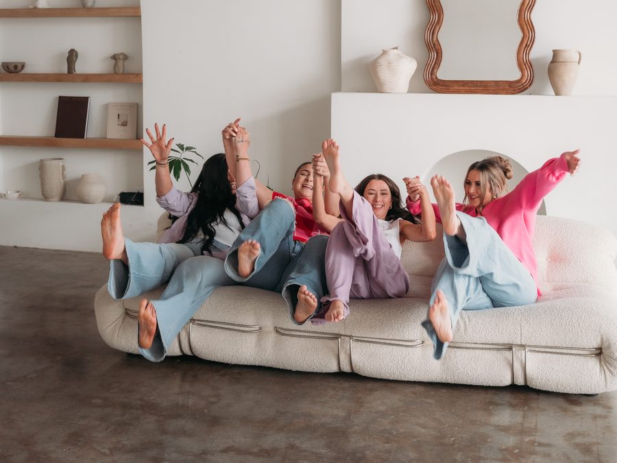 The four women of the Momeaze team on a sofa with their arms and feet in the air, laughing